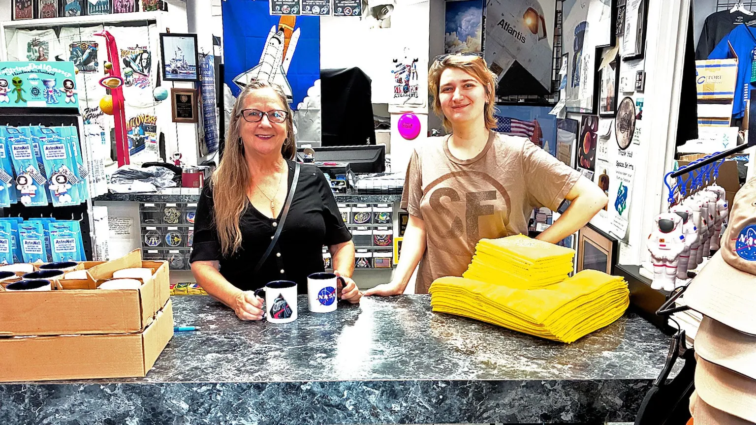 Pallab Ghosh/ Inside a small, crowded NASA gift shop, two people stand behind a grey marble-effect counter. Shelves and walls around them are packed with space souvenirs, mission posters, and astronaut photos. On the left are boxes of mugs; the woman holds two white mugs decorated with NASA-style logos. Next to her, a younger person in a pale T‑shirt leans on the counter near two neat piles of bright yellow folded T‑shirts. To the right, a rack displays beige and orange NASA baseball caps and small astronaut toys, giving the scene a busy, colourful, fan-filled atmosphere.