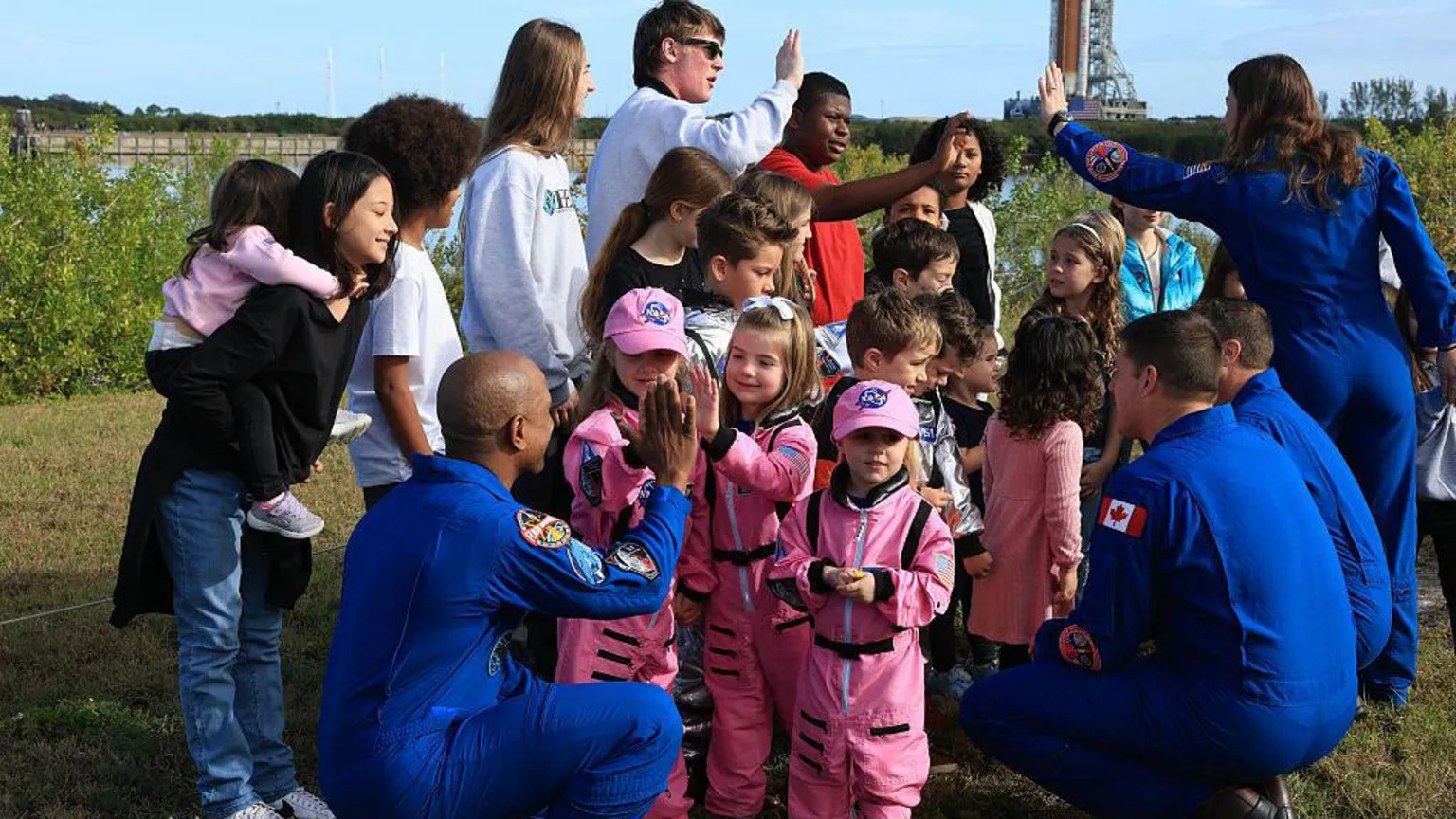 Joe Raedle/ On a grassy patch near water, three Artemis II astronauts in bright blue flight suits crouch down to talk with a group of young children. The children in the centre wear miniature pink spacesuits and caps, facing the astronauts and giving them high‑fives. Other children and parents cluster around them, some holding toddlers, forming a loose semicircle. In the distance, partly blurred, a tall rocket and launch tower rise above the trees. The mood is warm and playful, with astronauts and families smiling and interacting at eye level, turning a serious mission into a friendly, down‑to‑earth moment.