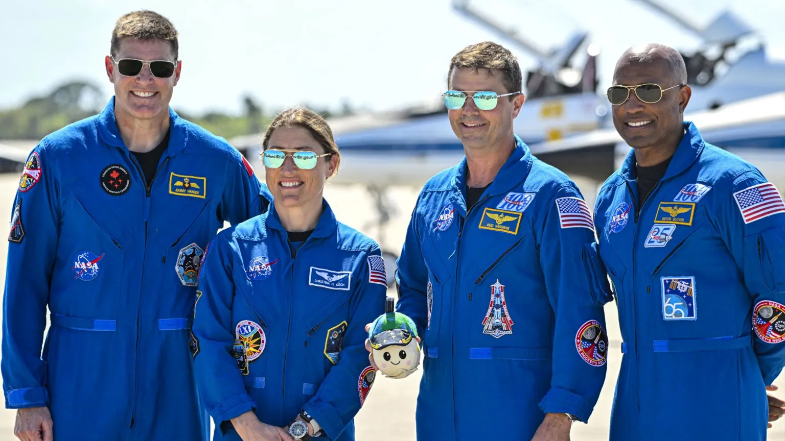 AFP via Four Artemis II astronauts stand side by side on a sunny runway, posing for a group portrait. They all wear bright blue NASA flight suits covered in mission patches and name badges, with dark boots. One astronaut in the centre holds a small mascot or model in both hands. Behind them, two sleek white-and-blue T‑38 training jets sit on the concrete, their pointed noses facing left and right, with the NASA “meatball” logo visible on a tail fin. The sky above is clear and pale blue, giving the scene a crisp, formal but upbeat feel.