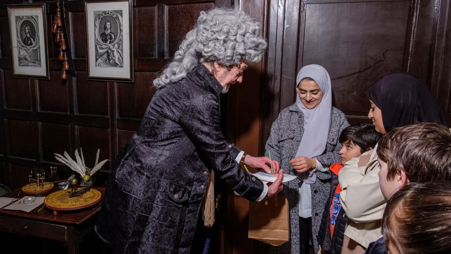 Bradford Museums/Phil Jackson A man in a grey curly wig and velvet coat speaks to some children and shows them some writing