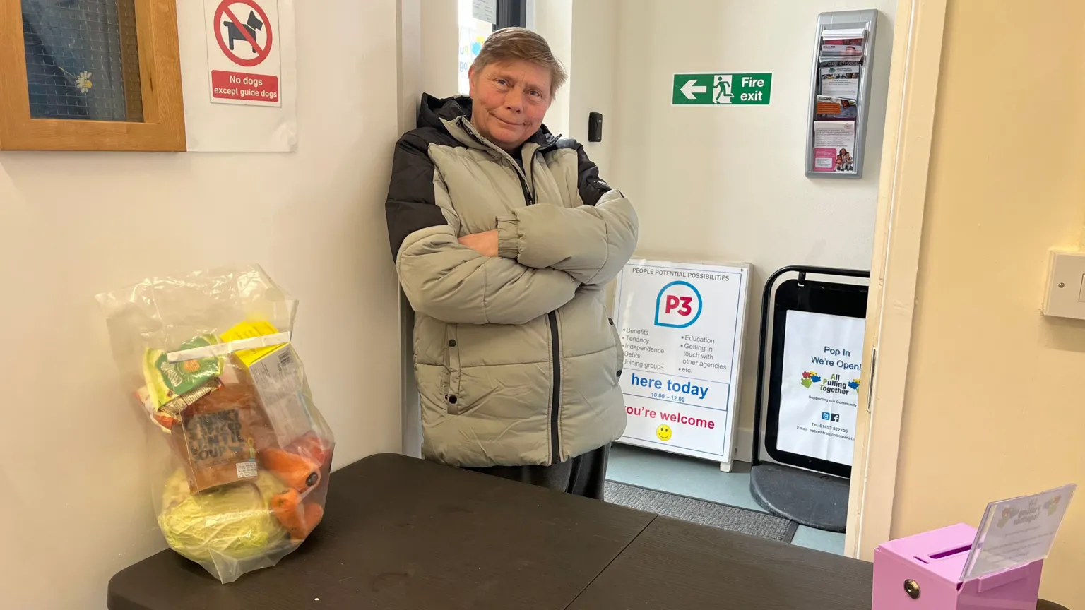 A woman wearing a grey puffer coat. She is smiling and has her arms folded. She's stood indoors, behind a table which has a bag of food on it.