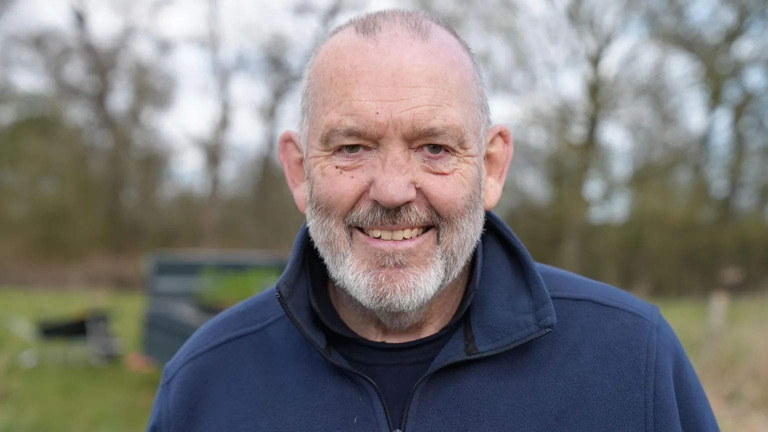 A man with a grey beard, and receding grey hairline is wearing a dark blue jacket. He is smiling and standing in front of blurred trees and grass.