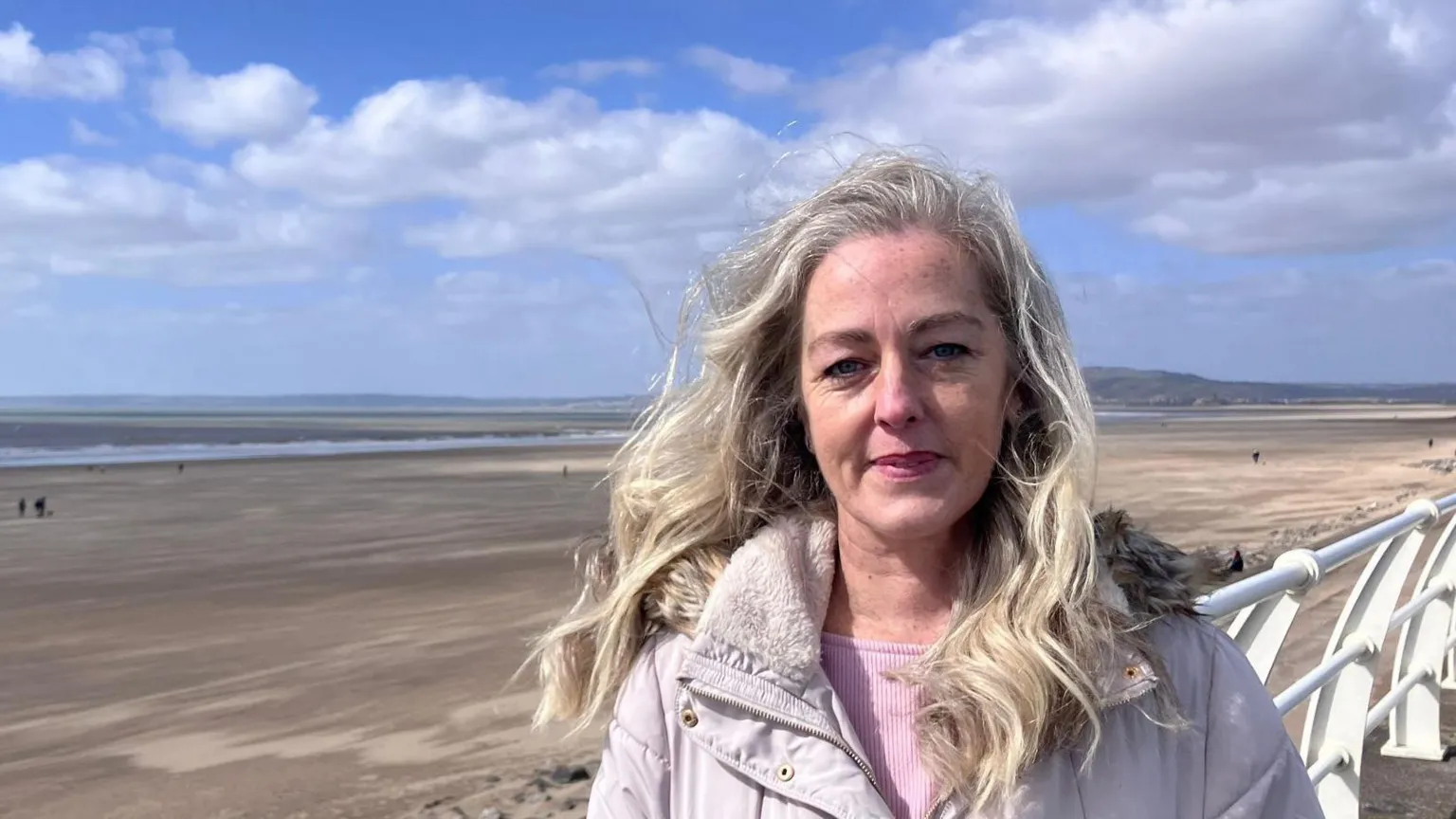 Jo Phillips smiles at the camera. She is standing on a promenade in front of a beach