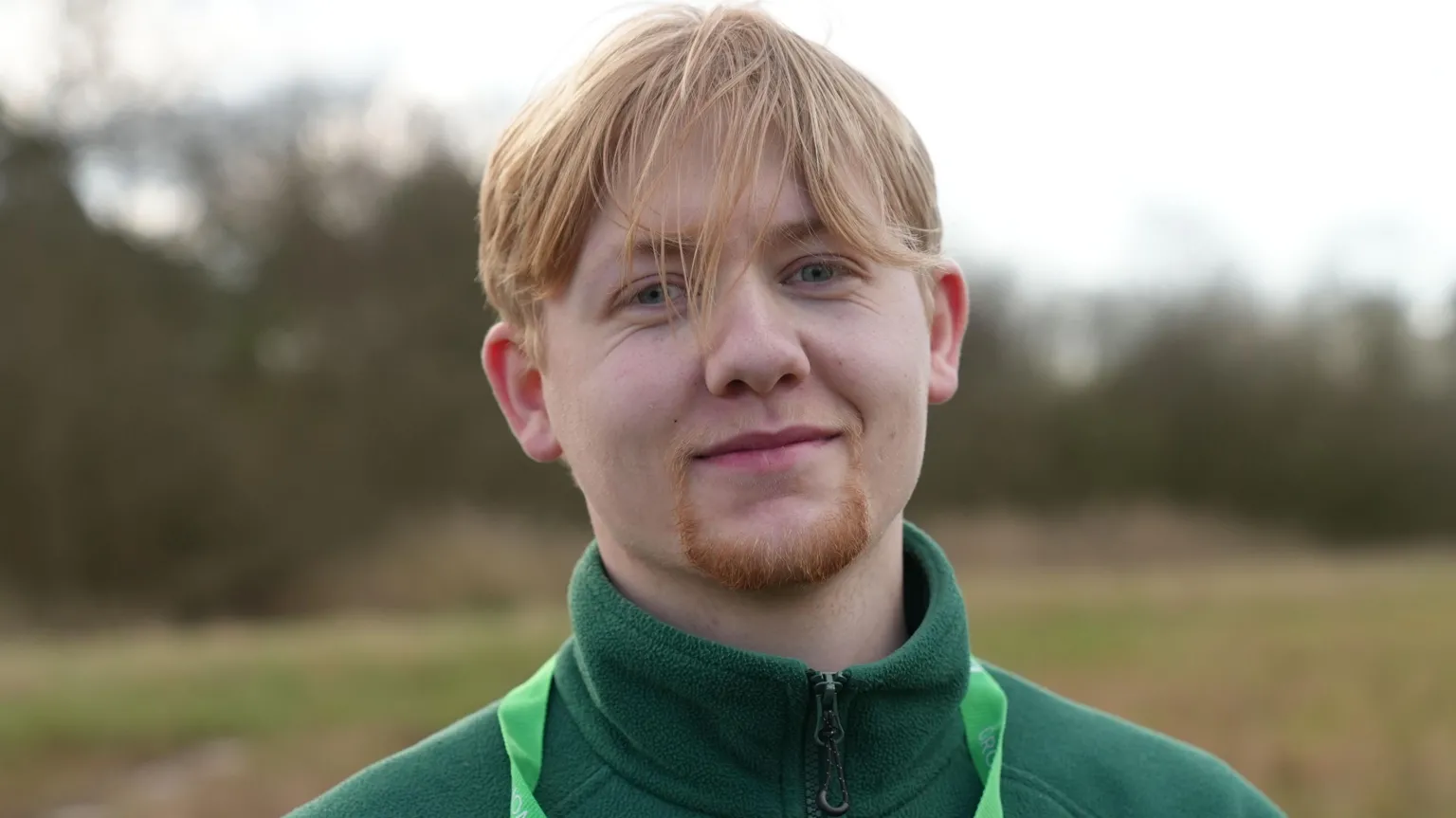 A man wearing a green outdoor jacket and a bright green lanyard is pictured standing in an open field, with blurred trees in the background. he has shortish blonde hair and a goatee beard.