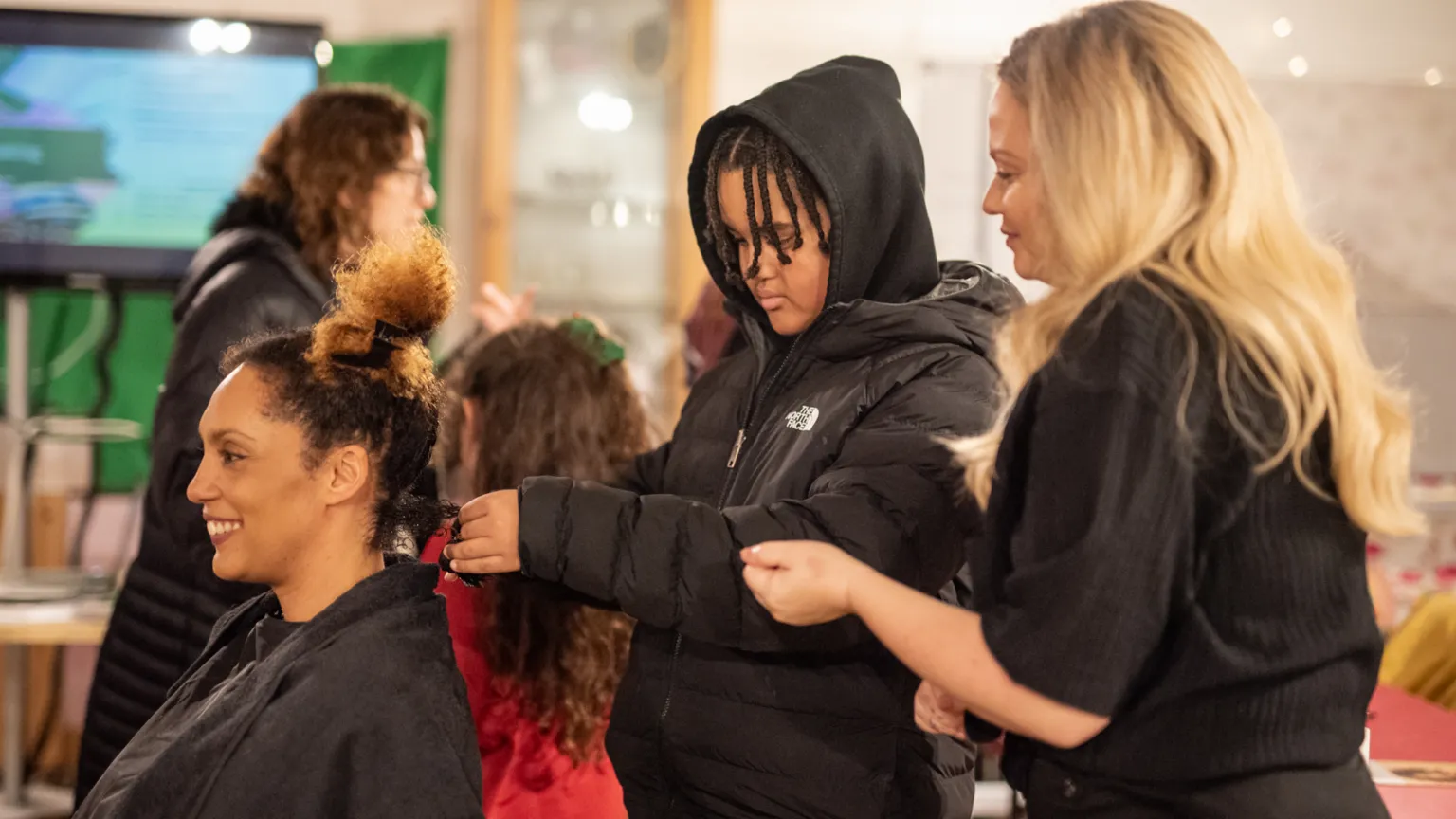 Marnie Bee Photography A woman with afro hair is smiling while sitting in a chair while a boy with braided afro hair is brushing her hair. A woman with long blonde hair is standing next to him and smiling while watching what he is doing.