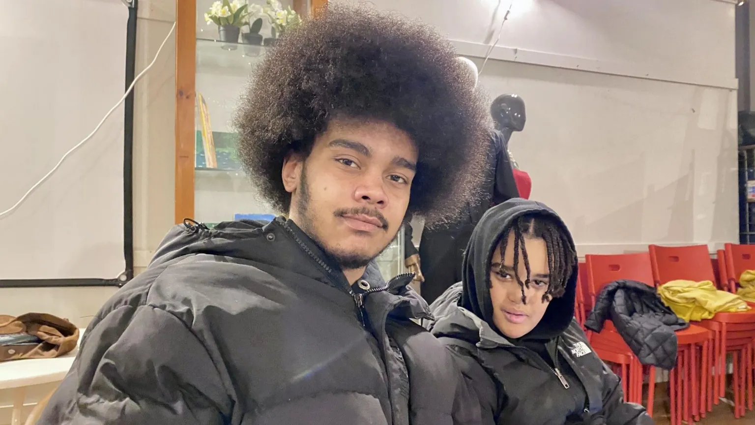 A young man and a boy are smiling while sitting next to each other at a table. The man is wearing a black coat and the boy is wearing a black coat and a black hooded top with the hood up.