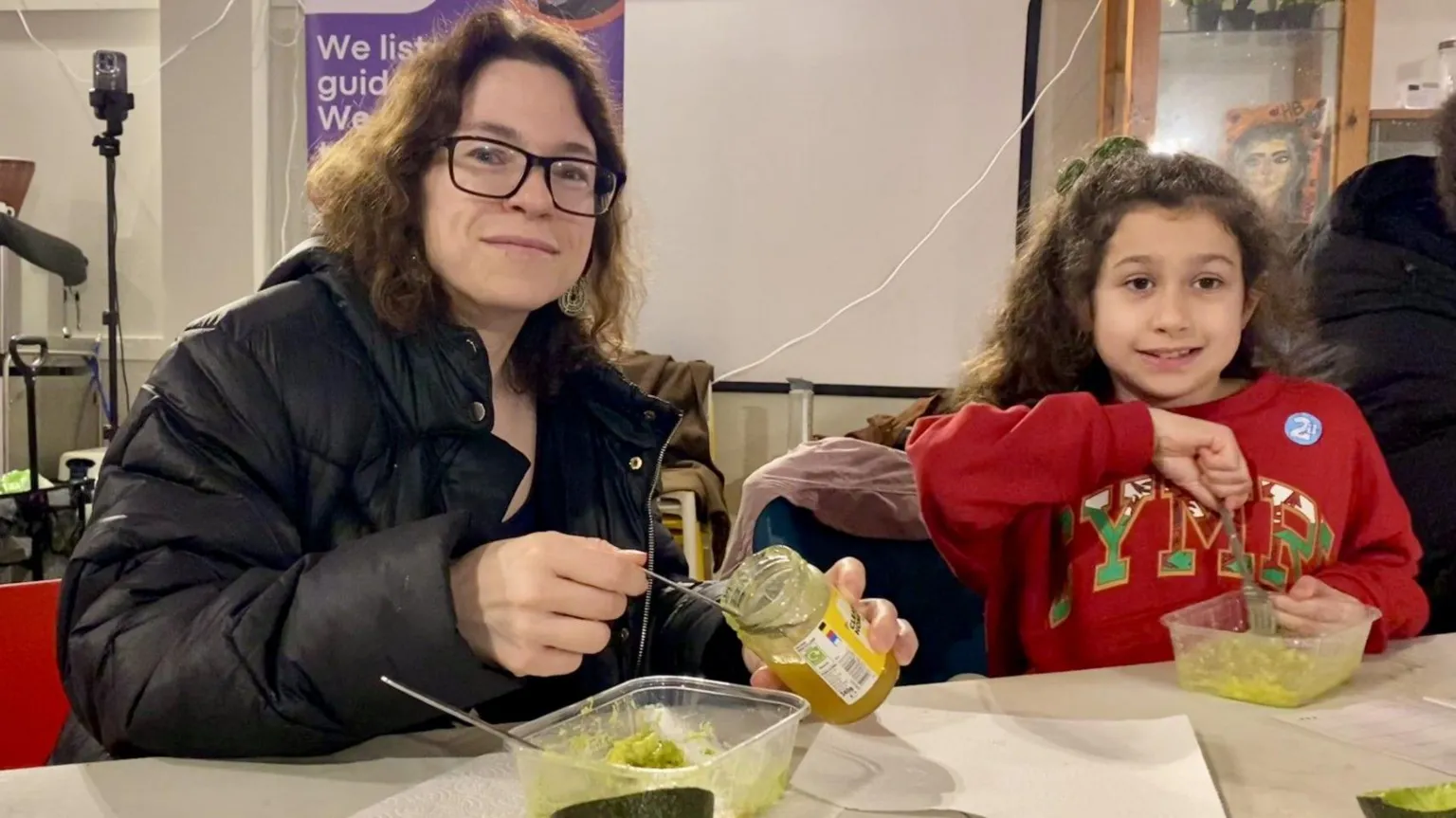 A woman with shoulder length curly brown hair is smiling as she holds a jar of honey. A plastic container with a green-coloured hair mask is on the table in front of her. A young girl with long curly brown hair and wearing a red sweatshirt with Cymru written on it is also smiling as she mixes a green-coloured hair mask in another plastic container.