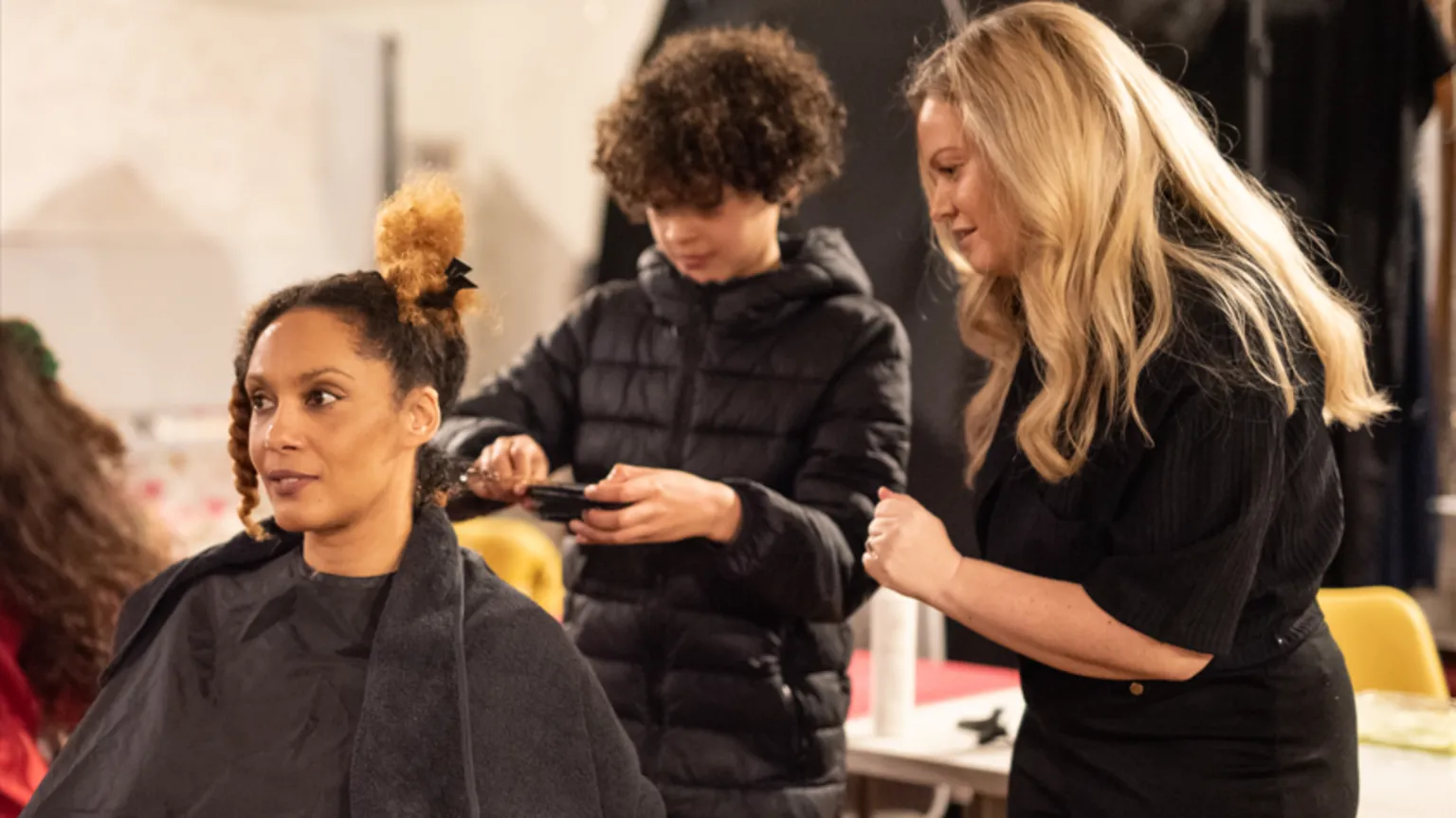 Marnie Bee Photography A woman with afro hair is smiling while sitting down with a black hairdressing gown on, as a young boy with afro hair is brushing her hair. A woman with long blonde hair is watching him.