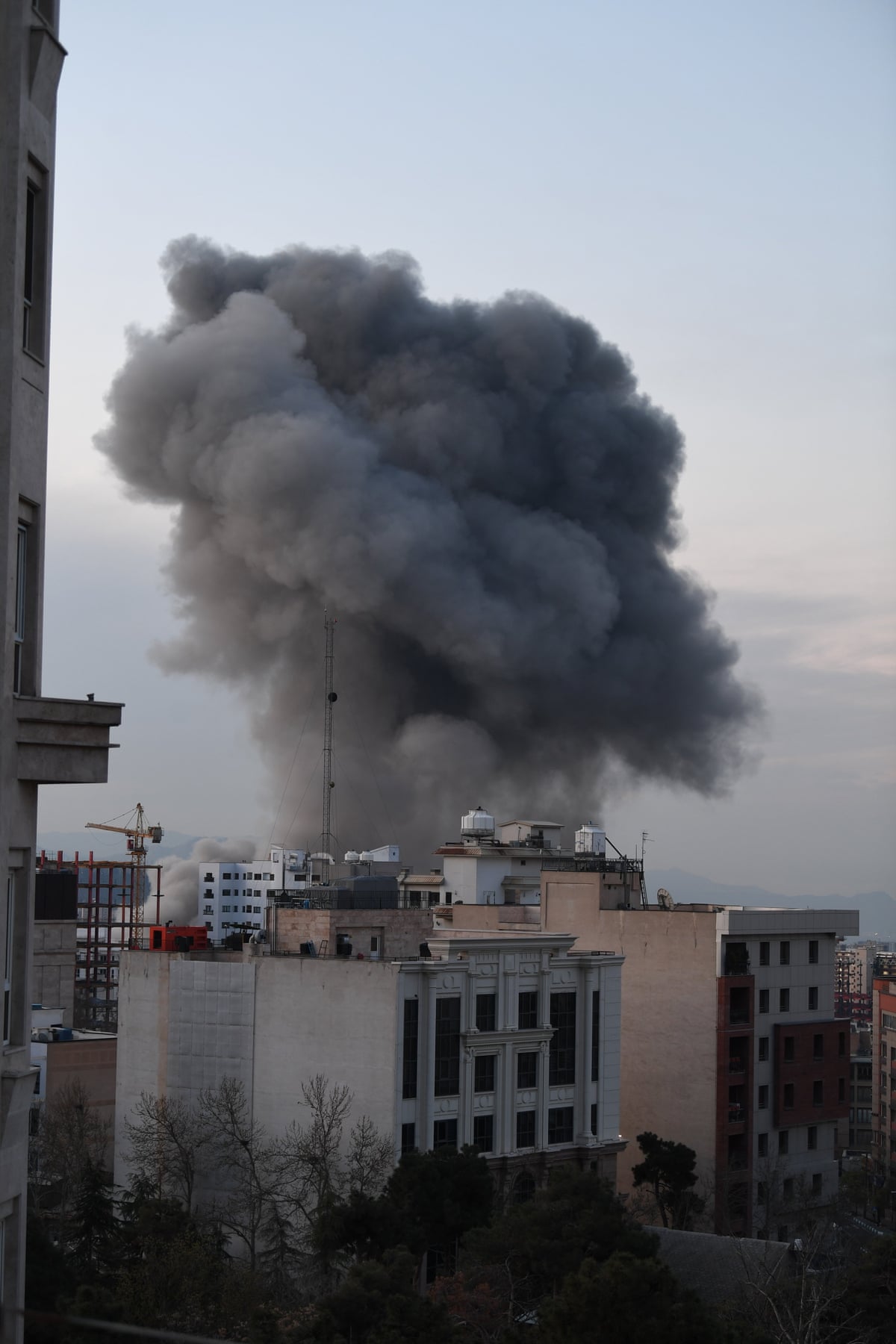 View of the city of Tehran with thick smoke rising into the air from behind a tall building.