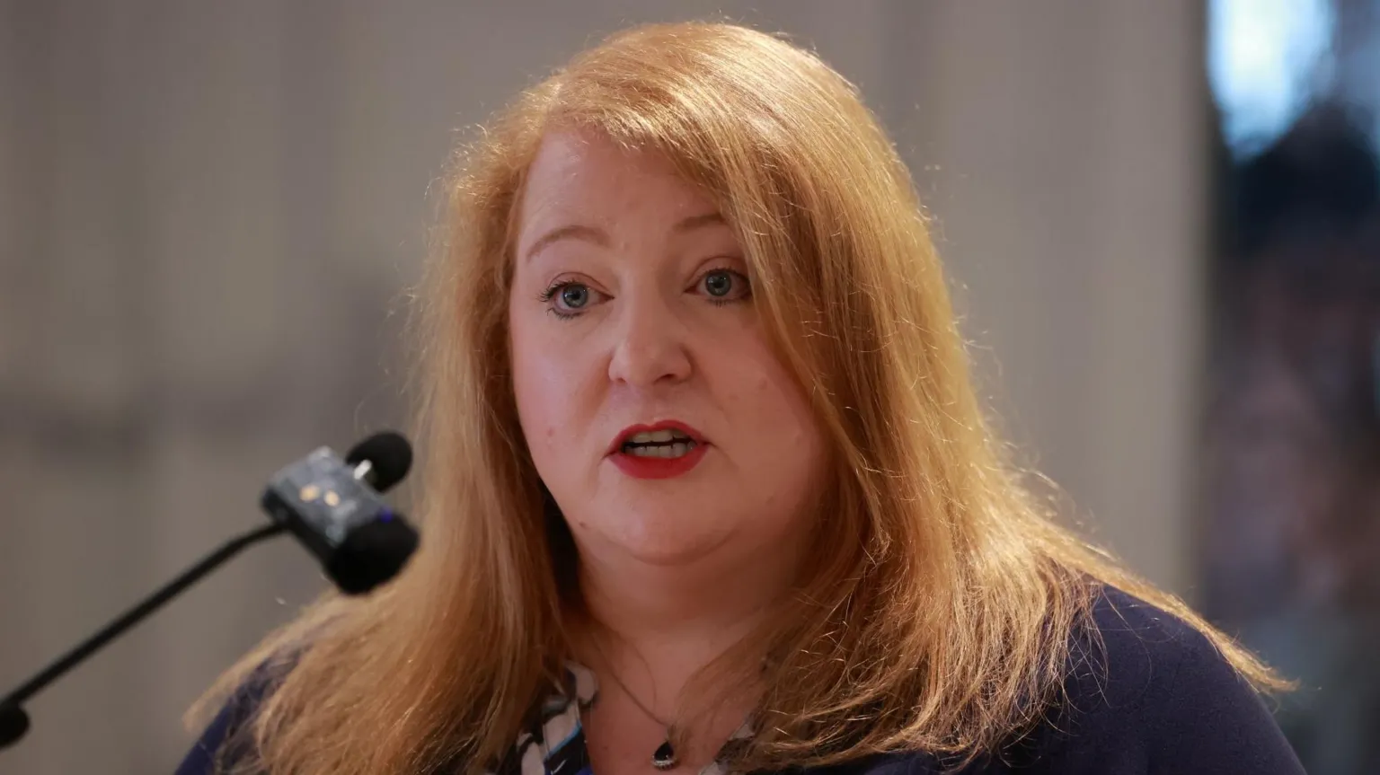 A close up image of Naomi Long. She has long, ginger hair. She is standing at a podium making a speech and has her mouth open. In front of her is a thin microphone with another wireless microphone attached.