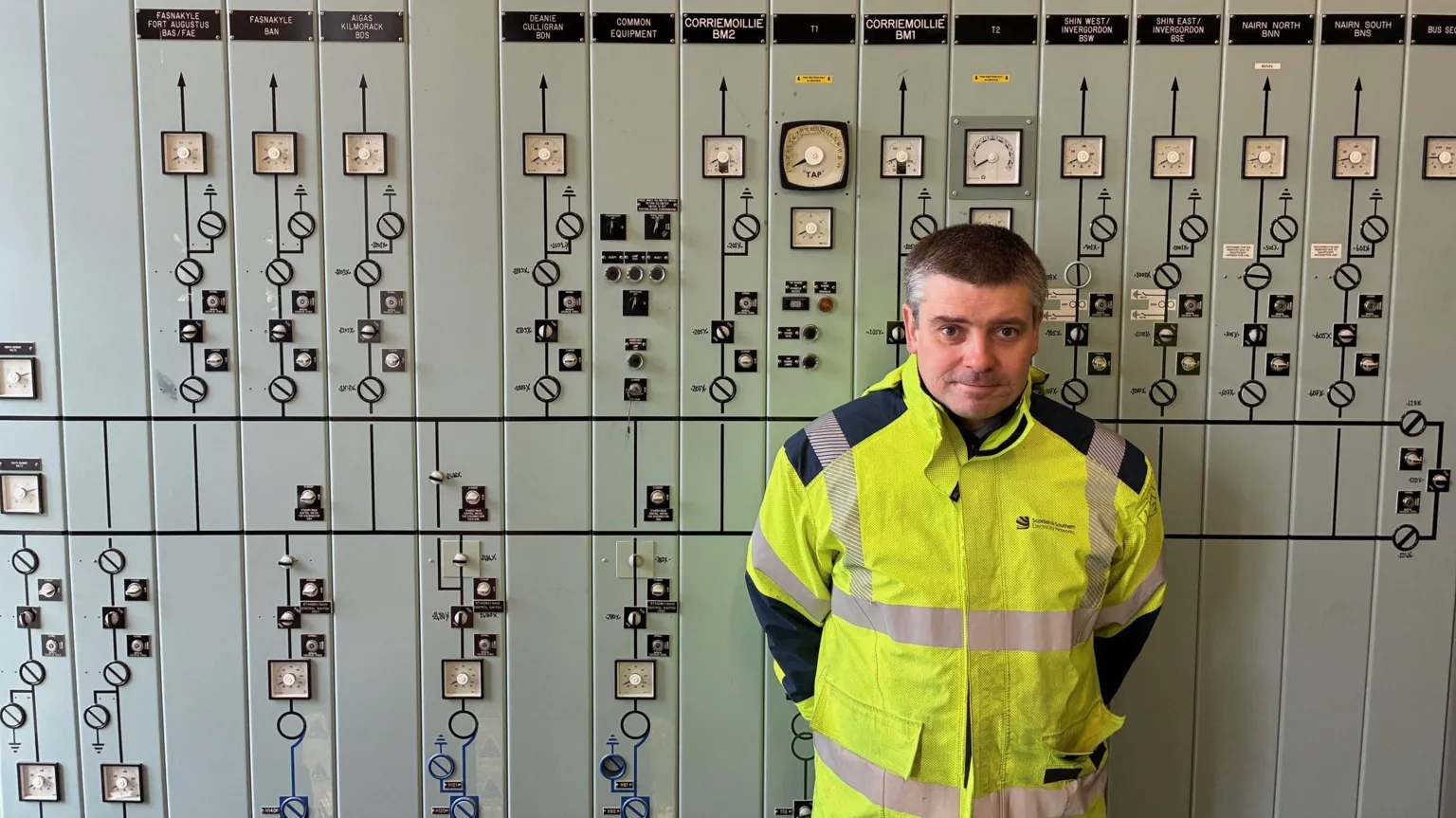 Greg Clarke wearing a high‑visibility yellow safety jacket stands in front of a large industrial control panel filled with dials, gauges, switches, and indicator lights.