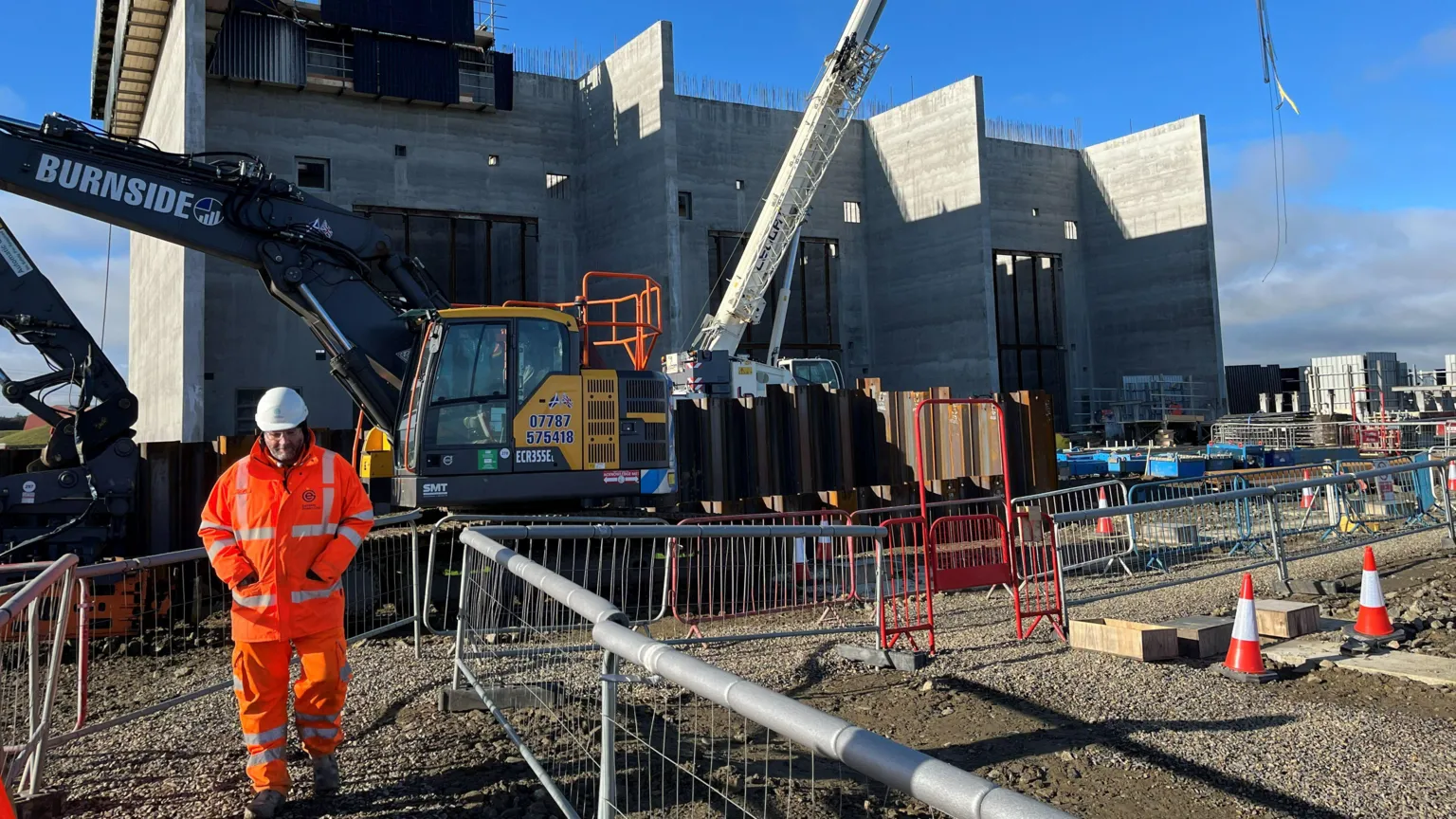 Peterhead building site, where the connector for the subsea cable is being built - Construction worker in high‑visibility orange gear walks across a large building site with heavy machinery, barriers, and a partially built concrete structure in the background.