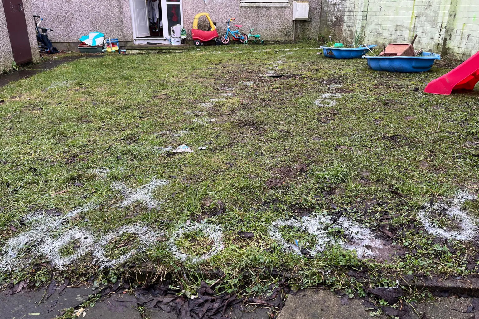 A general view of the back garden of the house at Ancrum Court. The grass has white writing spray painted on it containing letters and numbers.