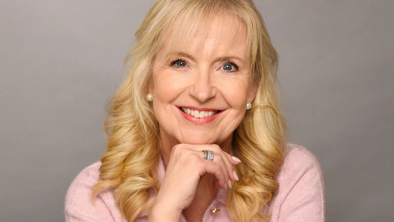 Carol Kirkwood smiling and resting her chin on her hand while wearing a pink top