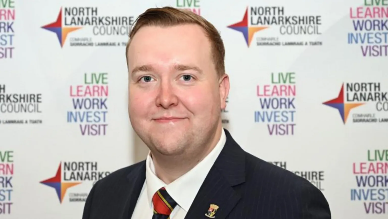 North Lanarkshire Council A man in a suit standing in front of a North Lanarkshire Council backdrop featuring repeated text promoting live, learn, work, invest, and visit