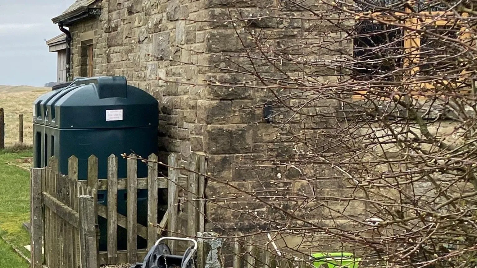 A tank sits in a garden next to a old house. The tank is large and green on located on the far side of a wooden fence 