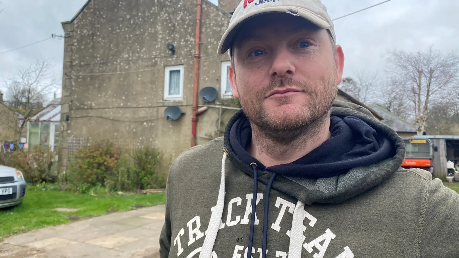 Richard Smith is a man in his 30s wearing a grey top with a cap and some stubble. He is standing in front of a large house with a conservatory just visible to the left