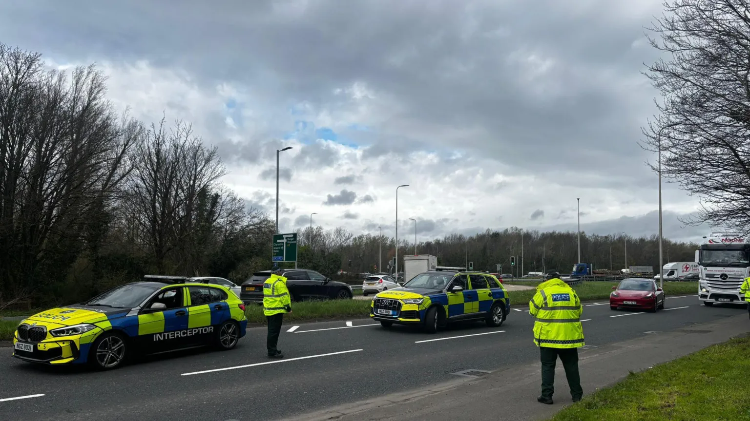 A road with two police cars parked in the middle. There are two police officers standing on the road, as well as a number of other vehicles. The sky is cloudy with patches of blue. 