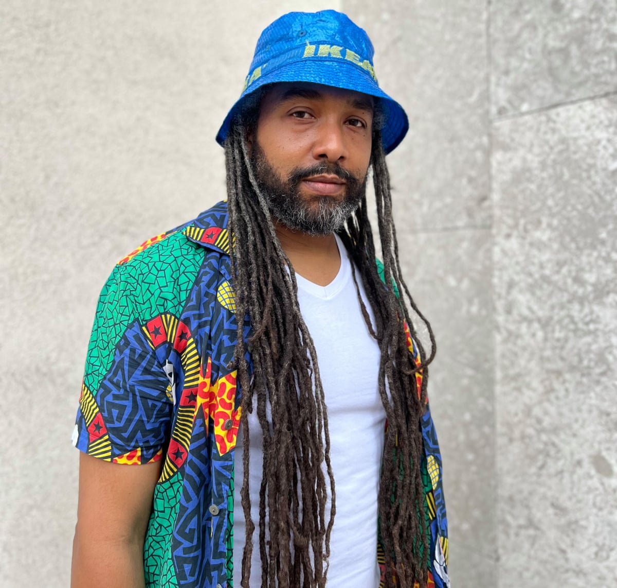 A man with dreads wears an Ikea bucket hat and colourful shirt and white vest.