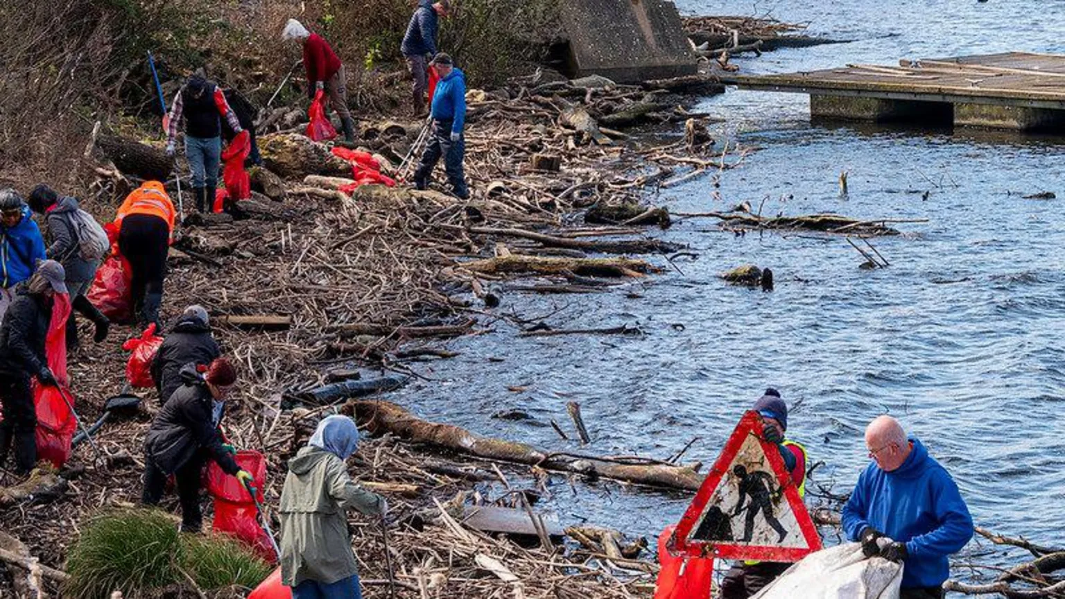 Cardiff Rivers Group Thirteen volunteers collecting litter into red plastic bin bags on the shore of Mermaid Quay in Cardiff Bay.