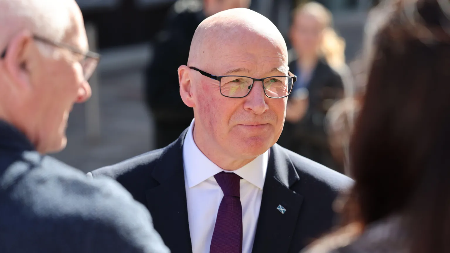 John Swinney, who is bald with glasses, wearing a business suit and a purple tie. He is facing two people, out of focus in the foreground
