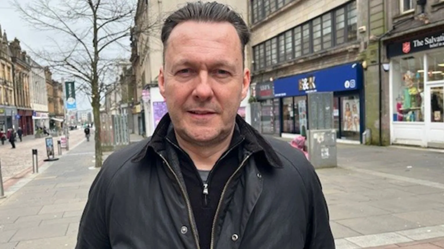 Russell Findlay, who has dark hair swept back, looks at the camera while sanding on a high street