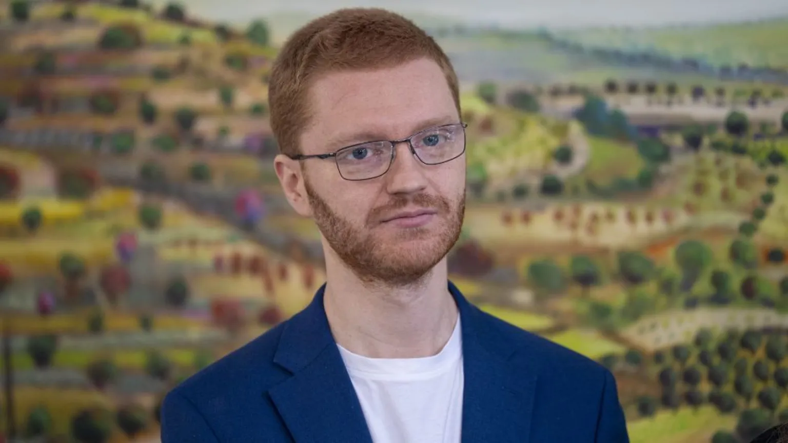 Ross Greer, who has short ginger hair and glasses, in front of an animated picture of green fields.