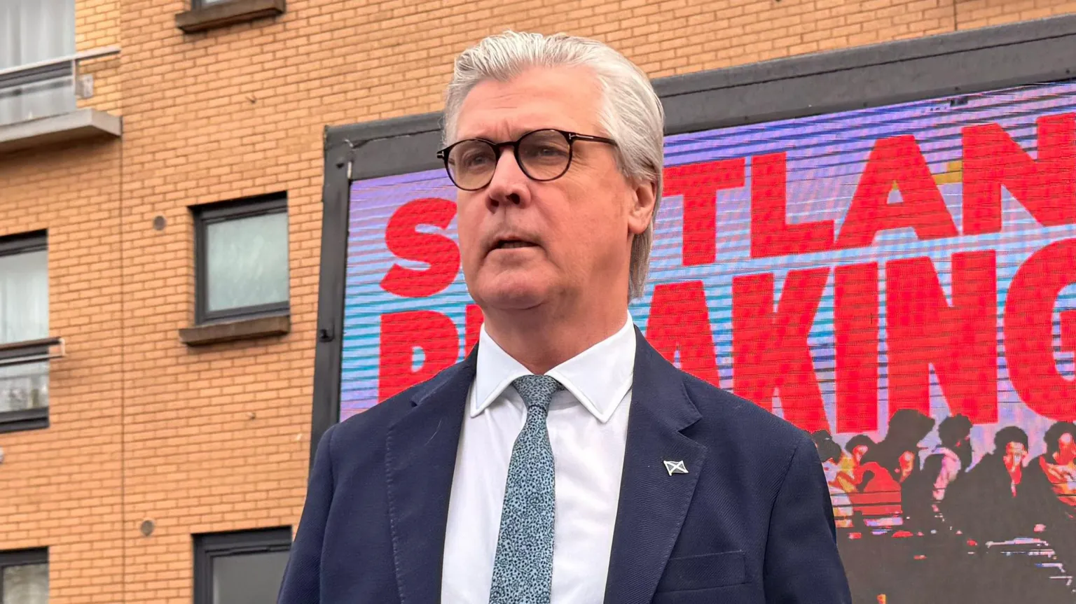 Malcolm Offord, who has grey hair and glasses, wearing a business suit while standing in front of a building and a digital billboard