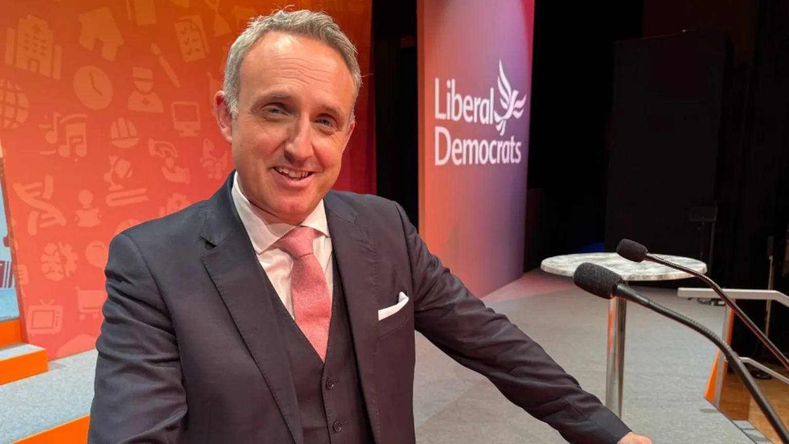 Alex Cole-Hamilton, who has short grey hair, poses at a podium with orange LibDem branding in the background