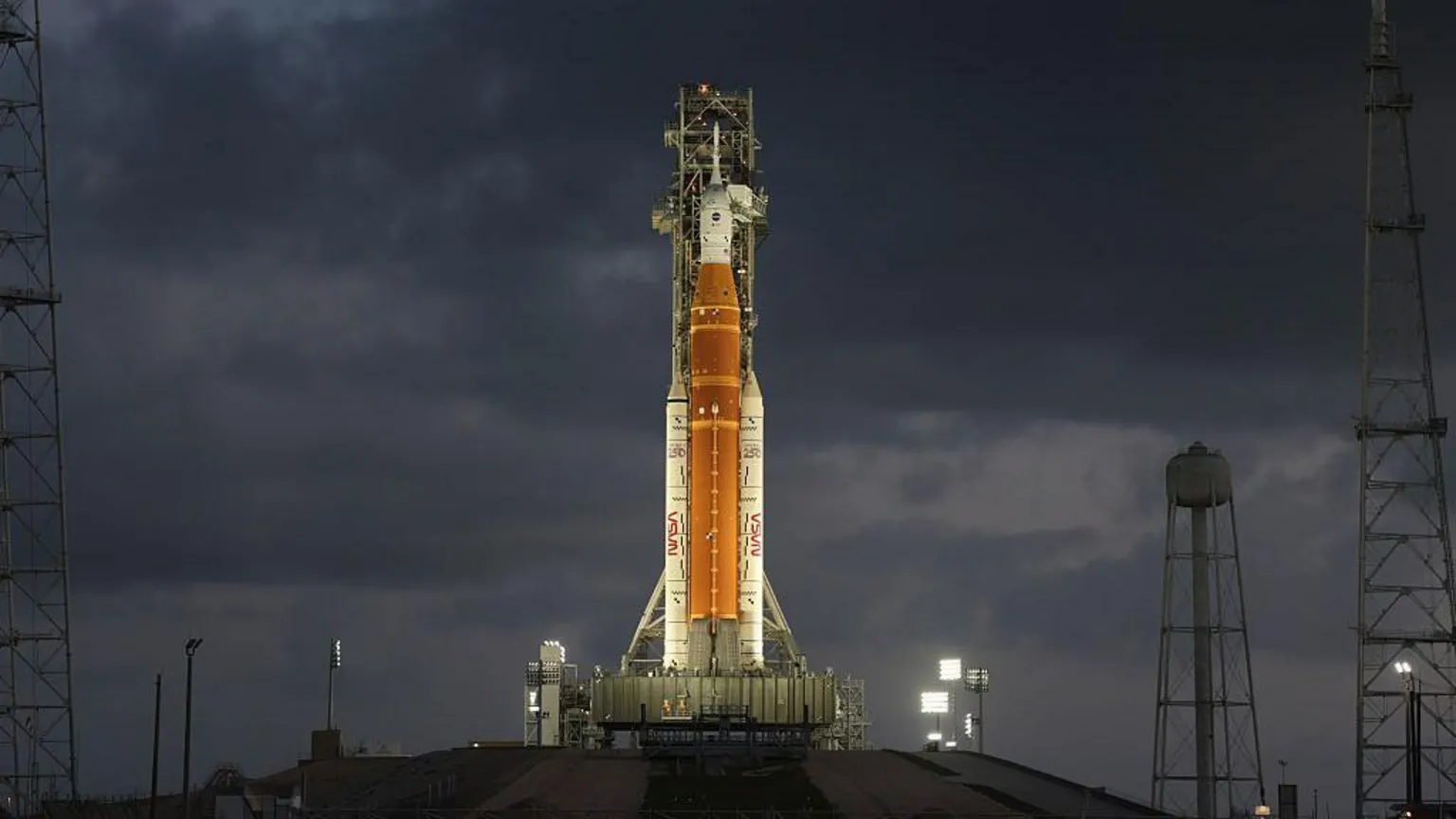  Orange rocket with white tip stands vertically ready to lift off lit up by flood lights.