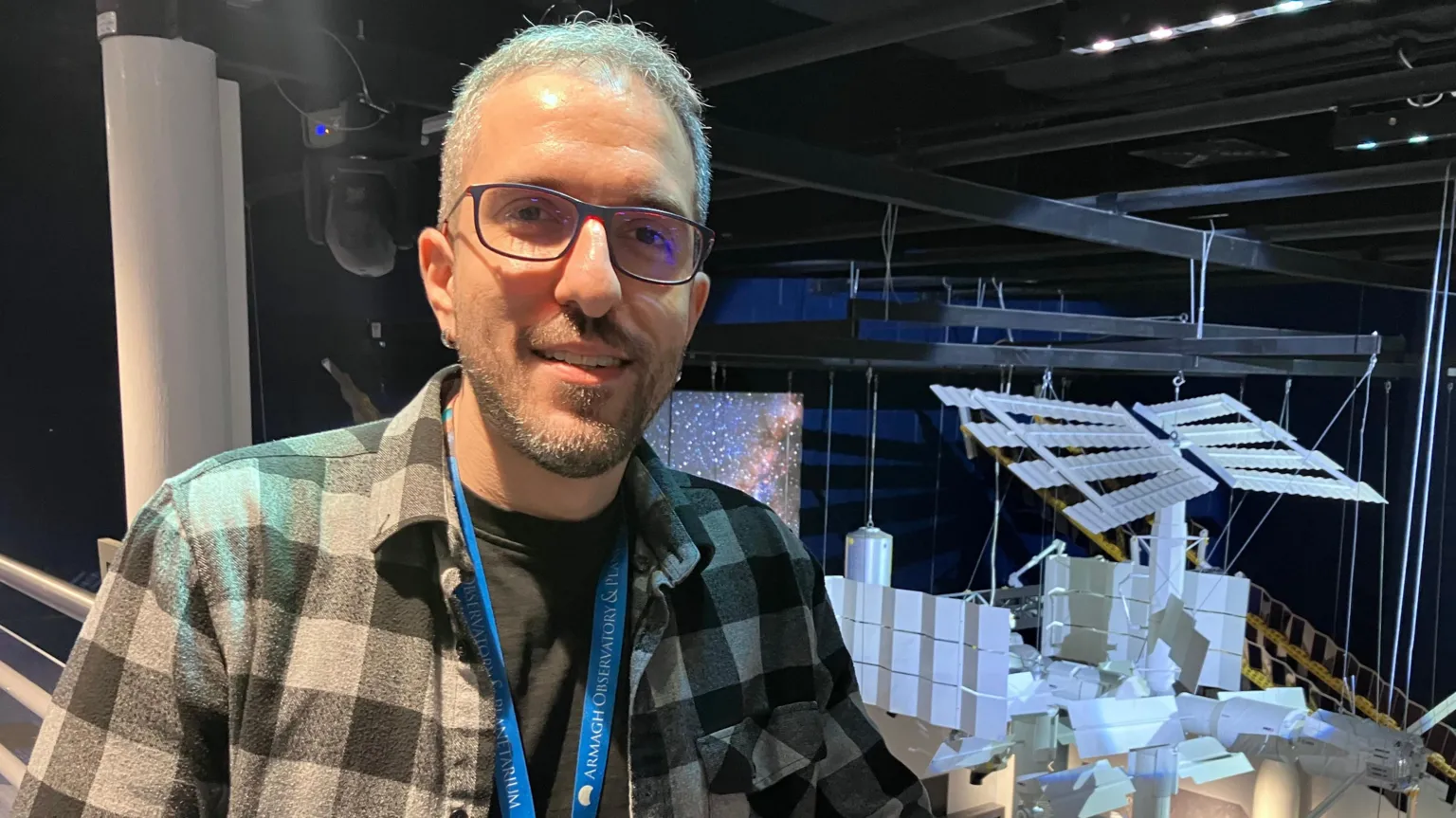 Kerem Osman Çubuk has short brown hair and glasses. He wears a grey and black chequered shirt with a black t-shirt underneath. Over his left shoulder is a white scaled-down model of the International Space Station. 