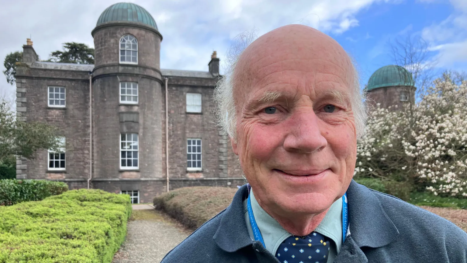 Michael Burton has thin grey hair. He wears a green shirt, navy tie and navy jumper. Behind him is an old grey Georgian building. With seven clear glass windows across it. Between the man and the building are low green hedges which run either side of a stone patch. 