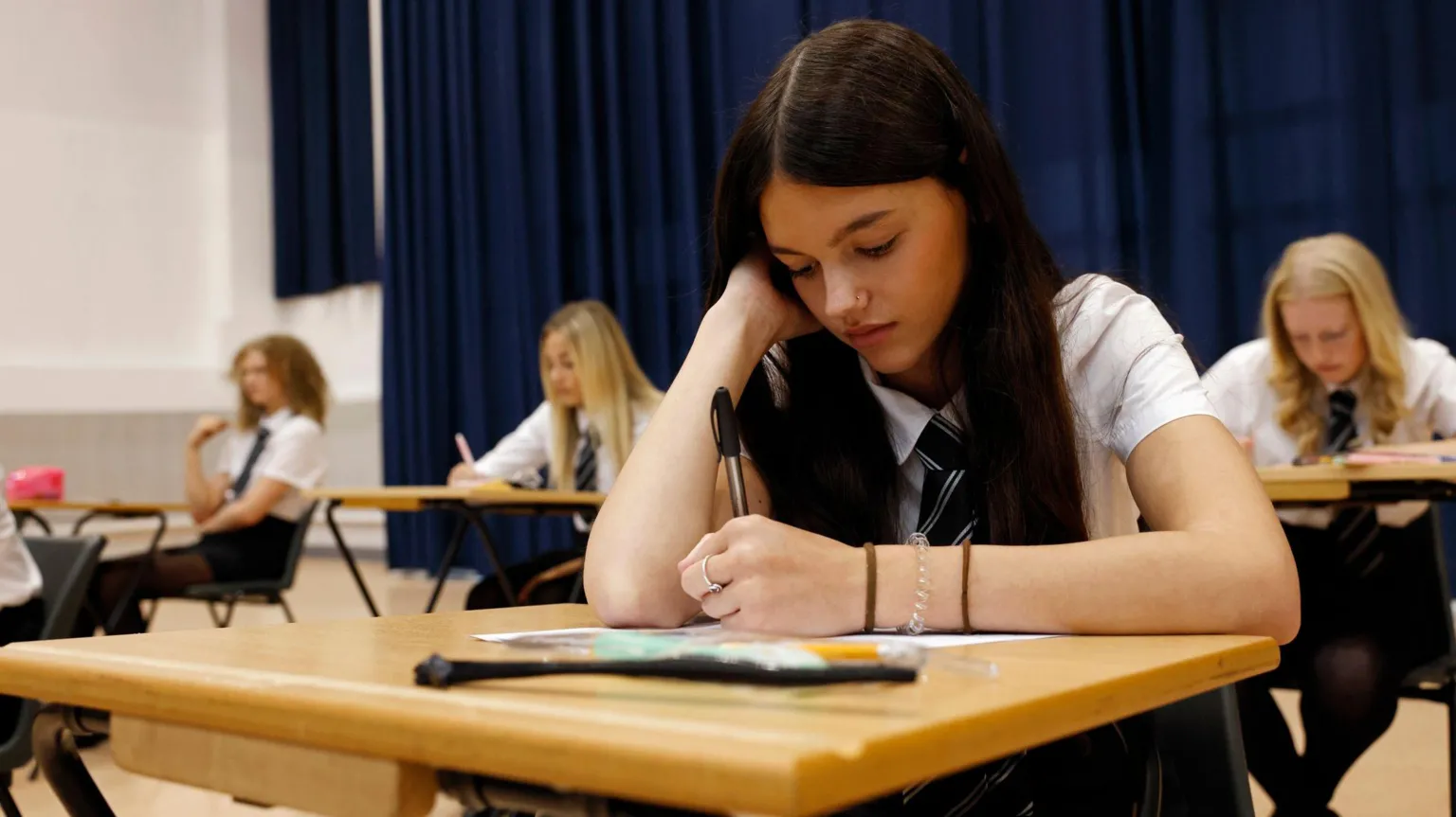  A female secondary school student sits an exam in a large school hall concentrating on her paper. She has long brown hair and is wearing a white shirt and a school tie.