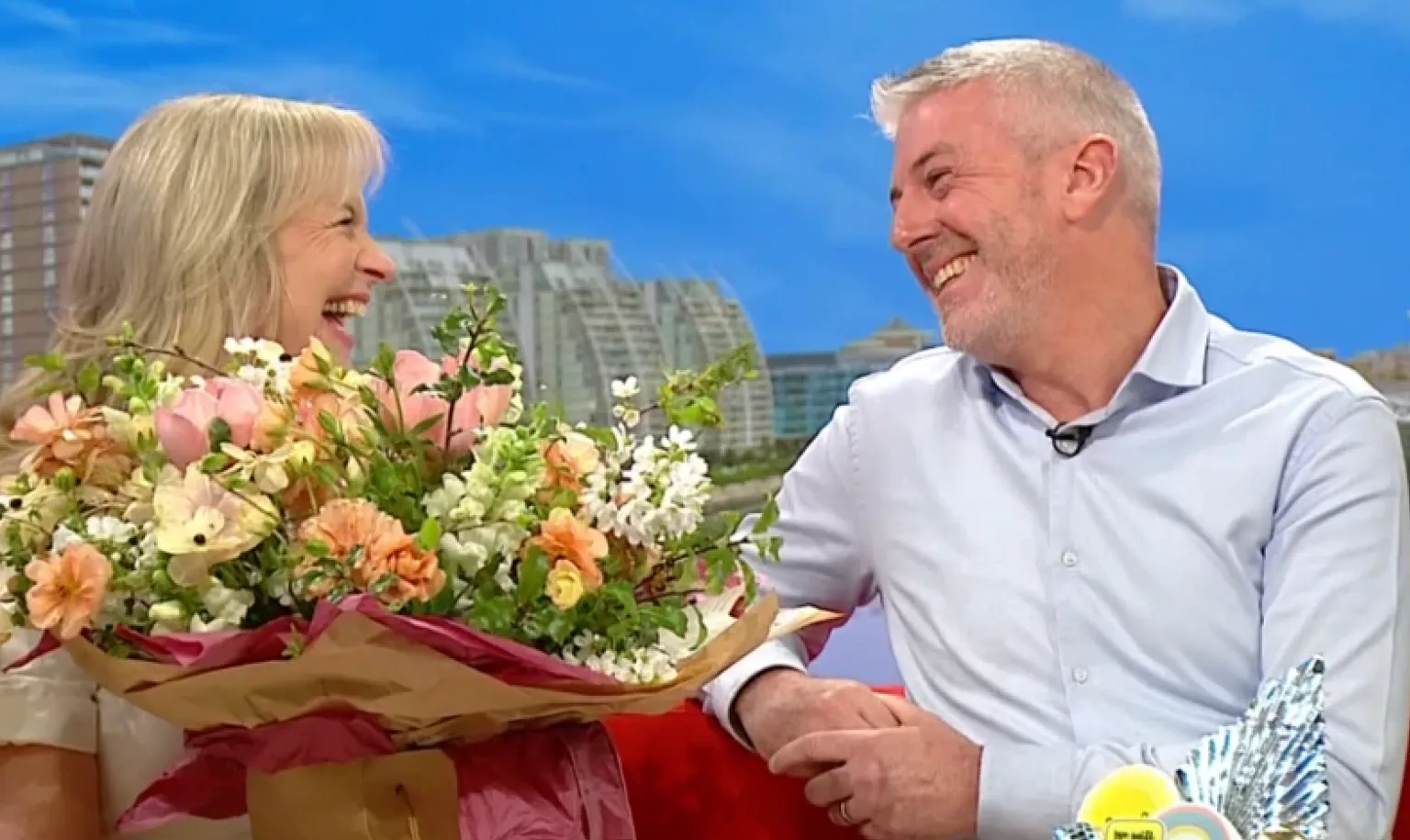 Carol Kirkwood sits on the red BBC Breakfast sofa with a bouqet of flowers next to her husband Steve. They both look at each other and smile.