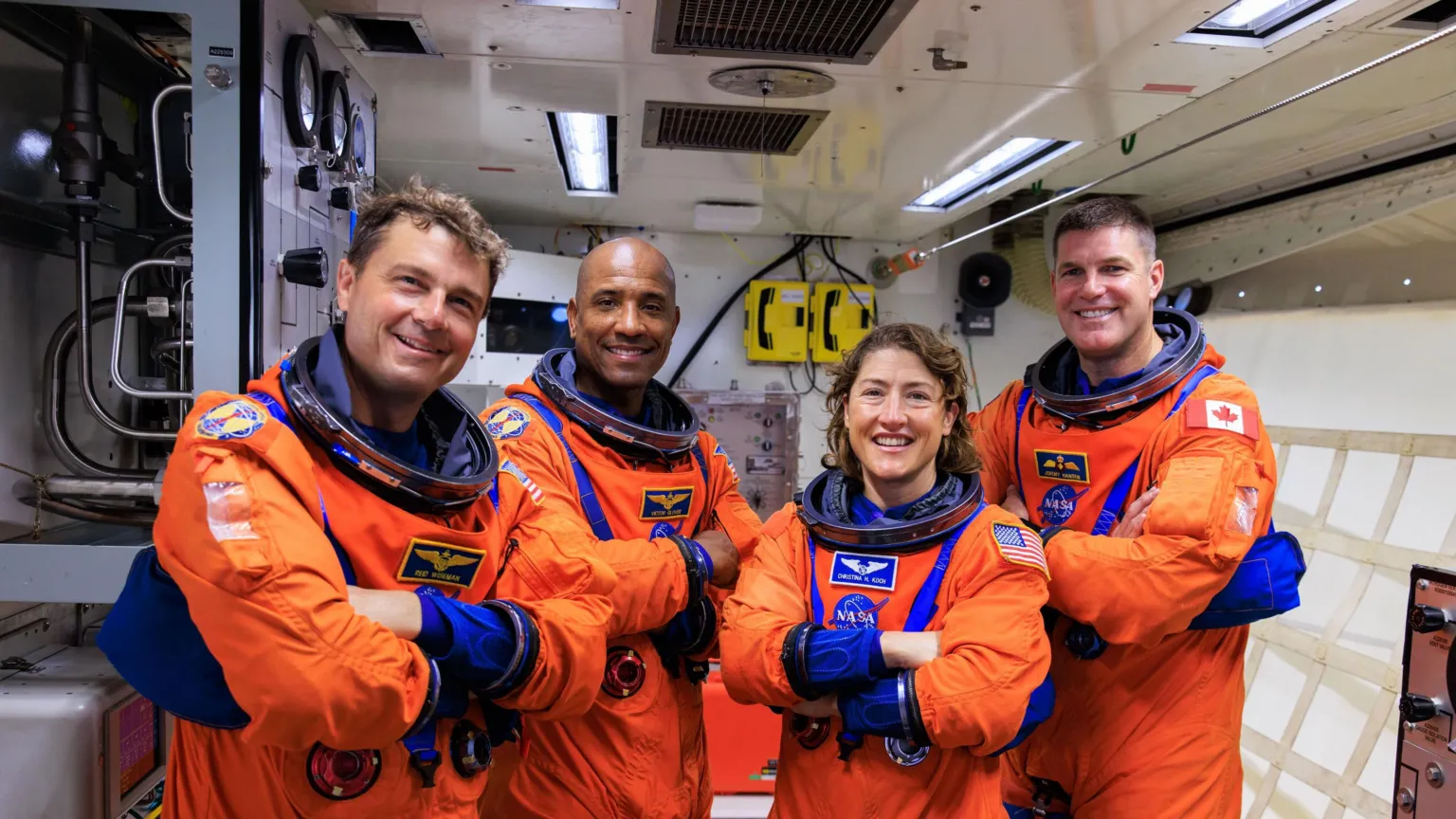 NASA Four astronauts stand side by side inside a cramped white spacecraft or support module, wearing bright orange launch and entry suits with blue trim and mission patches. Their arms are folded confidently across their chests.