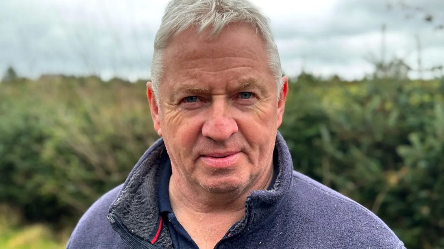 a man in a fleece looking at the camera. Behind him a hedge is visible