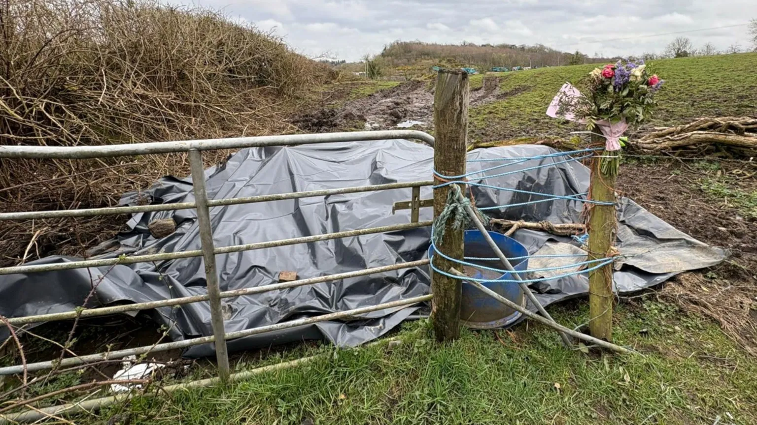 a large tarpaulin behind a fence. A bunch of flowers is tied to the fence post
