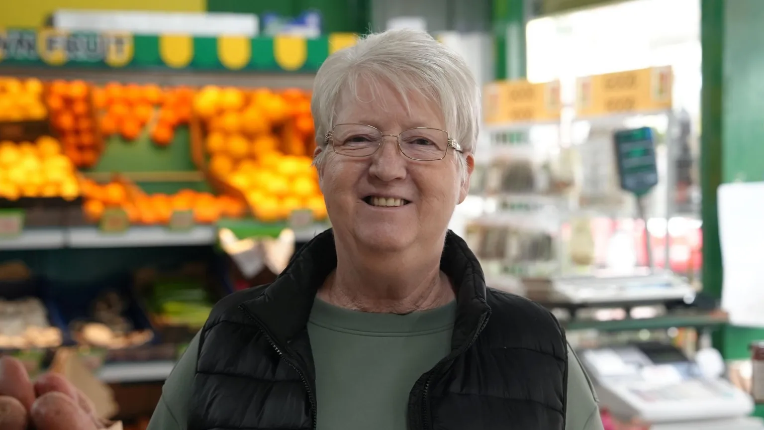 Barbara Farmer has short grey hair and glasses. She is wearing a green top and a black gillet and standing in a greengrocers. 