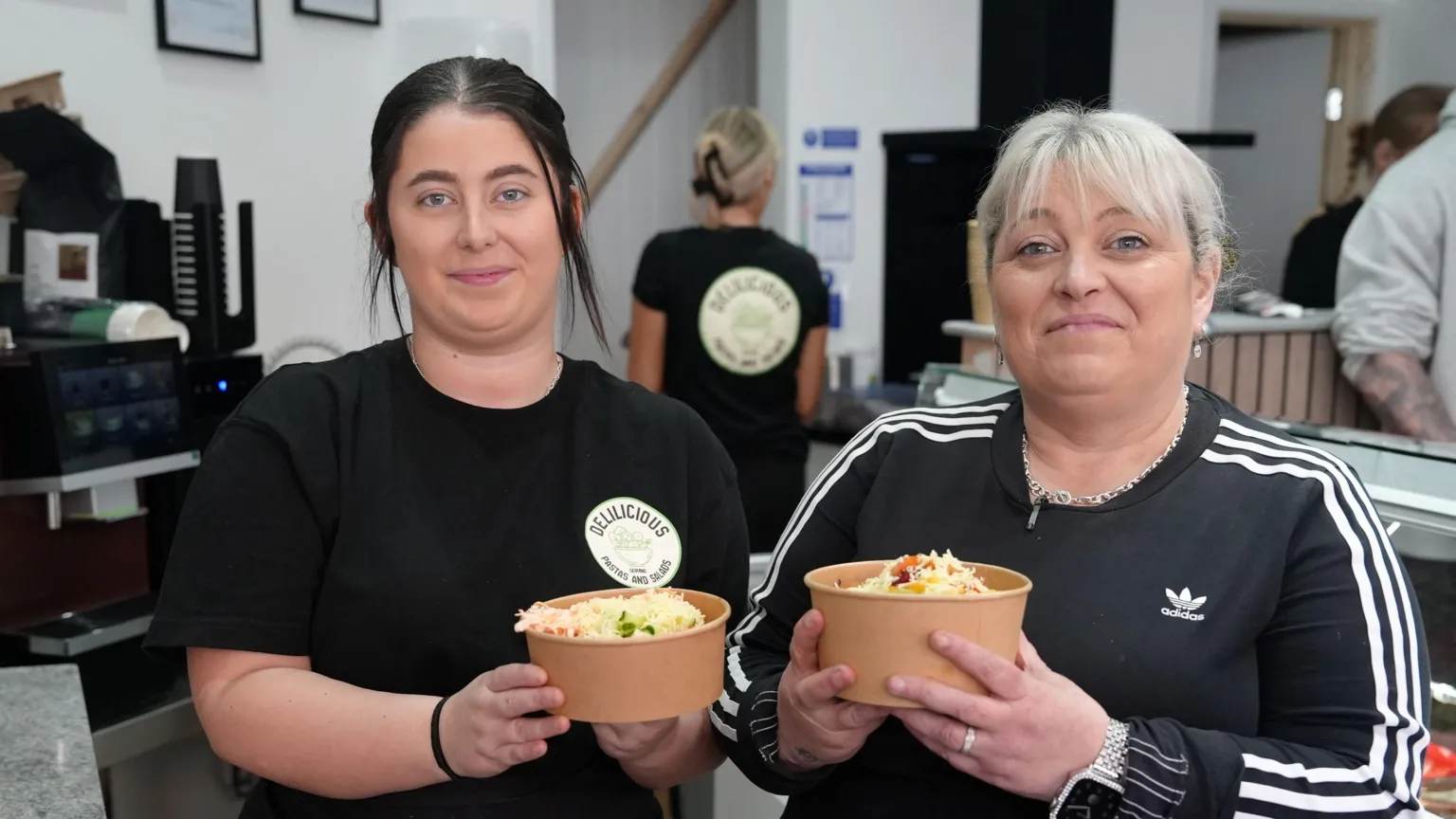 Joanne and Charlie Bruford are standing behind the counter of their new pasta and salad bar holiday a salad box each. Charlie has dark hair toed off her face, Joanne has blonde hair with a fringe tied off her face. Both are wearing black. 