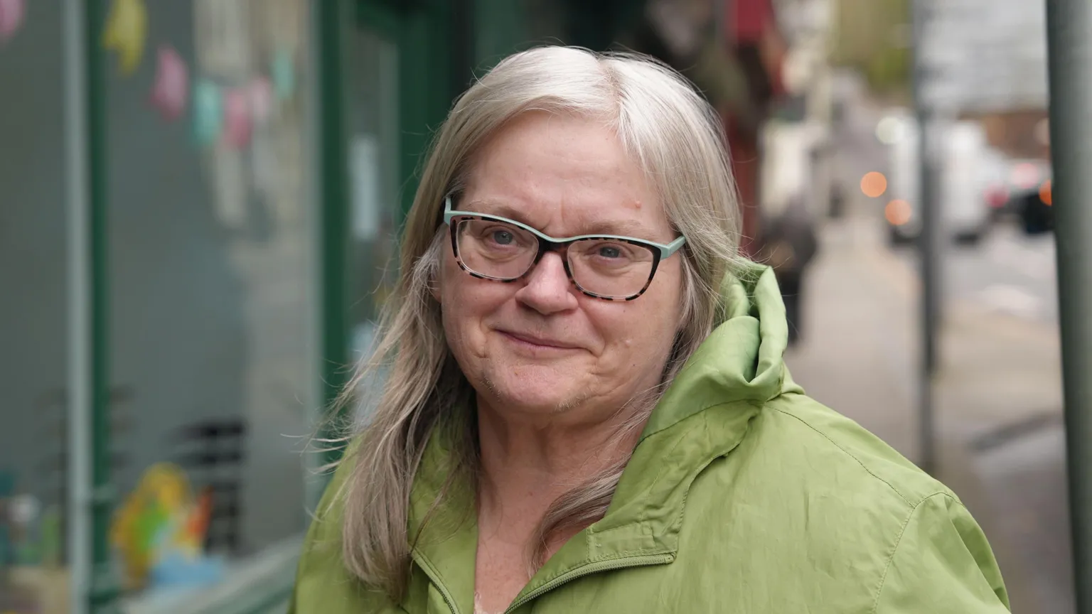 Julie Evans has long fair hair and is wearing glasses and a green jacket. She is standing on a street.