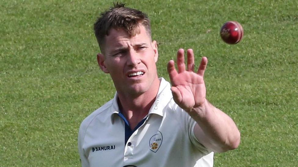 Luis Reece, wearing Derbyshire's white jersey with the club badge on, catches a ball with an outstretched left arm in preparation to bowl a delivery