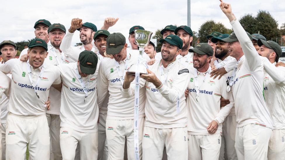 Leicestershire players celebrate in a large group together as Ian Holland lifts the trophy aloft in the centre to after winning promotion from Division Two