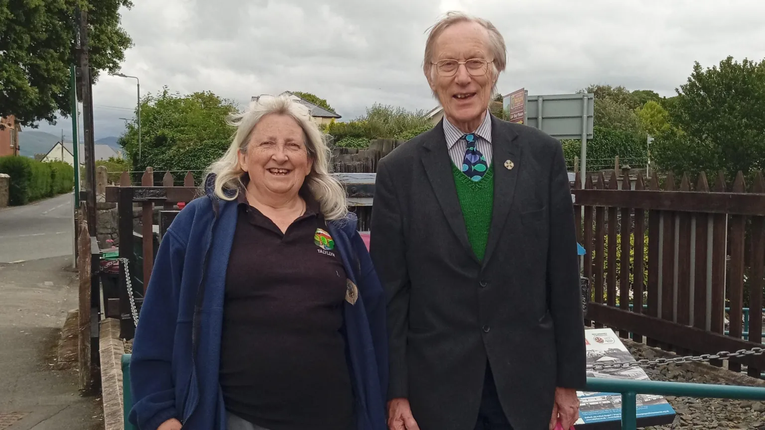Diocese of Bangor Susan and Roger stood outside standing next to each other. Susan has long grey hair and glasses on top of her head. She is wearing a black polo shirt and navy blue jacket. Roger has short grey hair. He is wearing glasses, a dark grey blazer, green vest, striped shirt and tie with blue, green and purple fish on it.