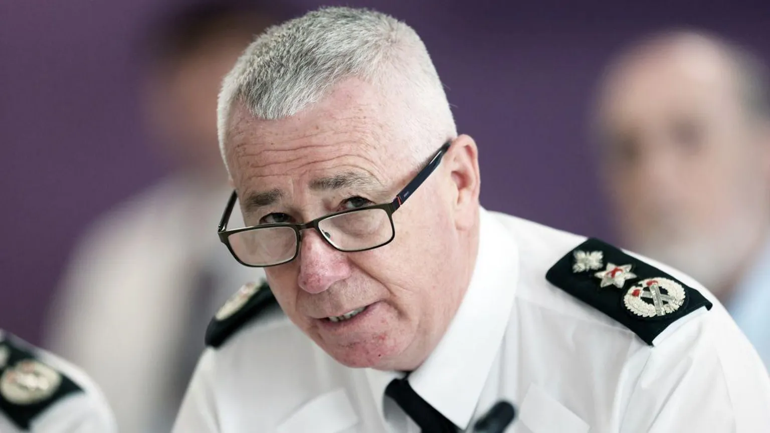  Jon Boutcher has light grey hair and is wearing black-framed glasses. He is wearing his police shirt and a black tie