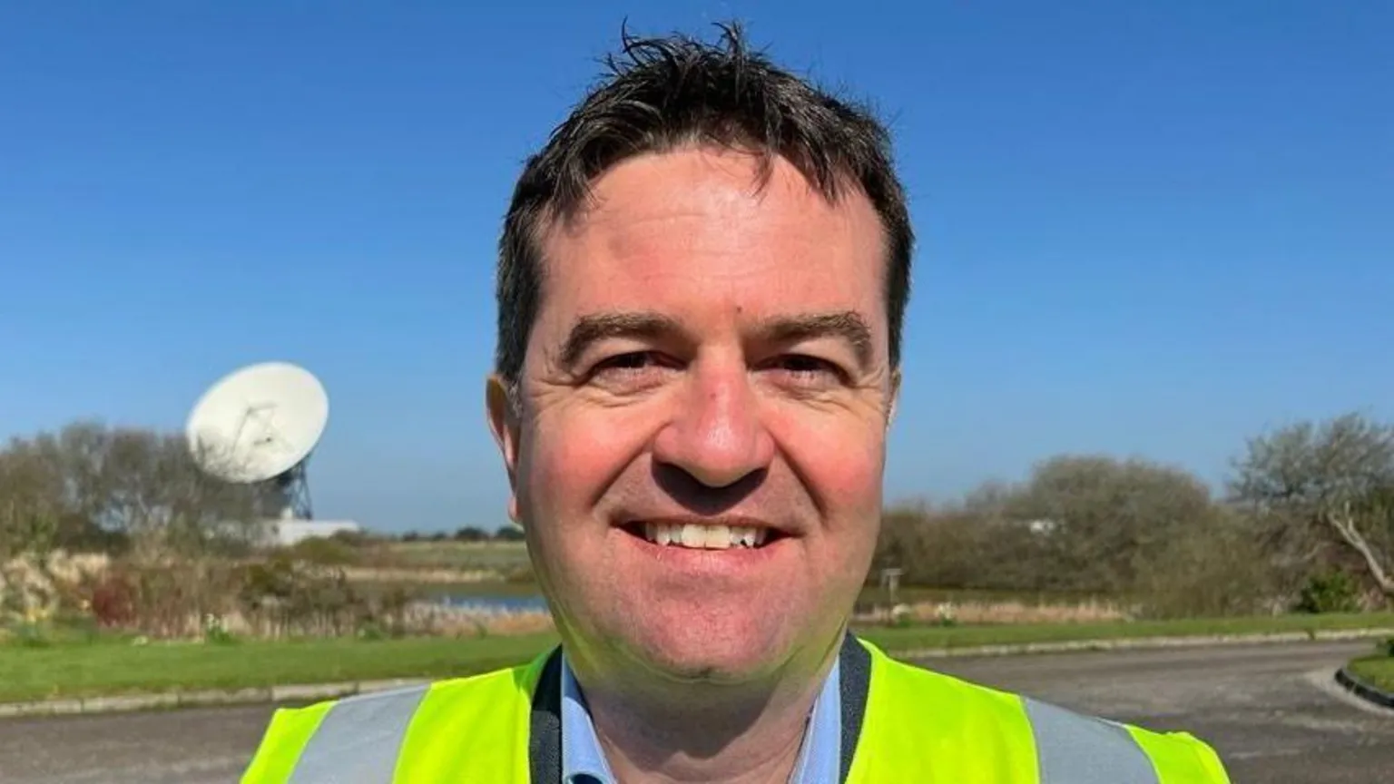 A picture of Matthew Cosby - he is wearing a blue suit shirt, black blazer and hi-visibility jacket. Behind him is a satellite dish.