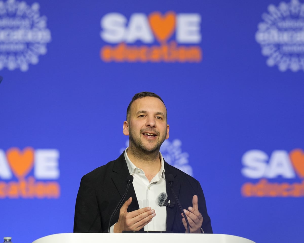 Zack Polanski speaks on stage in front of branding, saying 'save education'