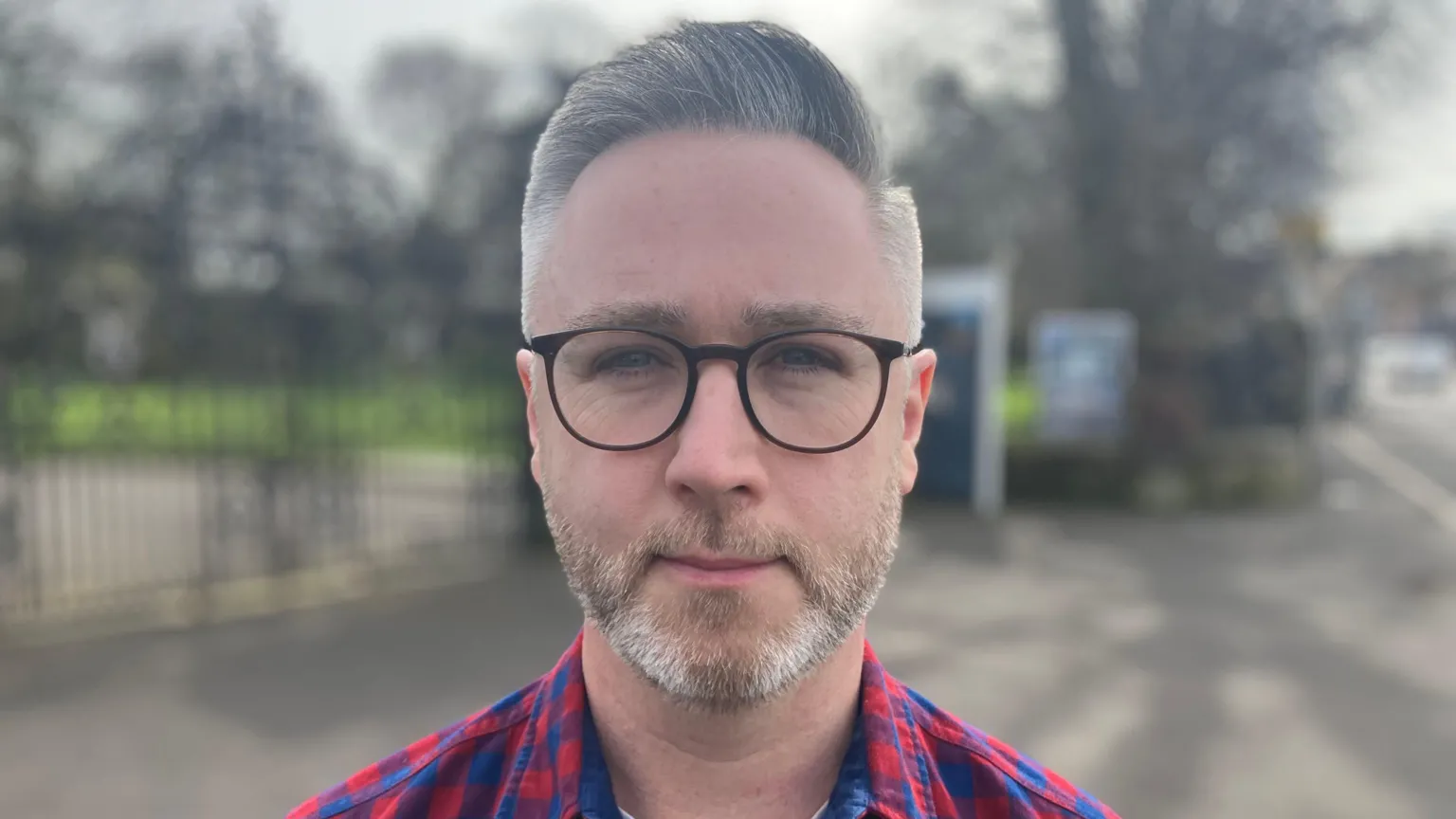 A man looks directly at the camera. He is wearing glasses and a red plaids shirt. He has grey hair and light stubble. 