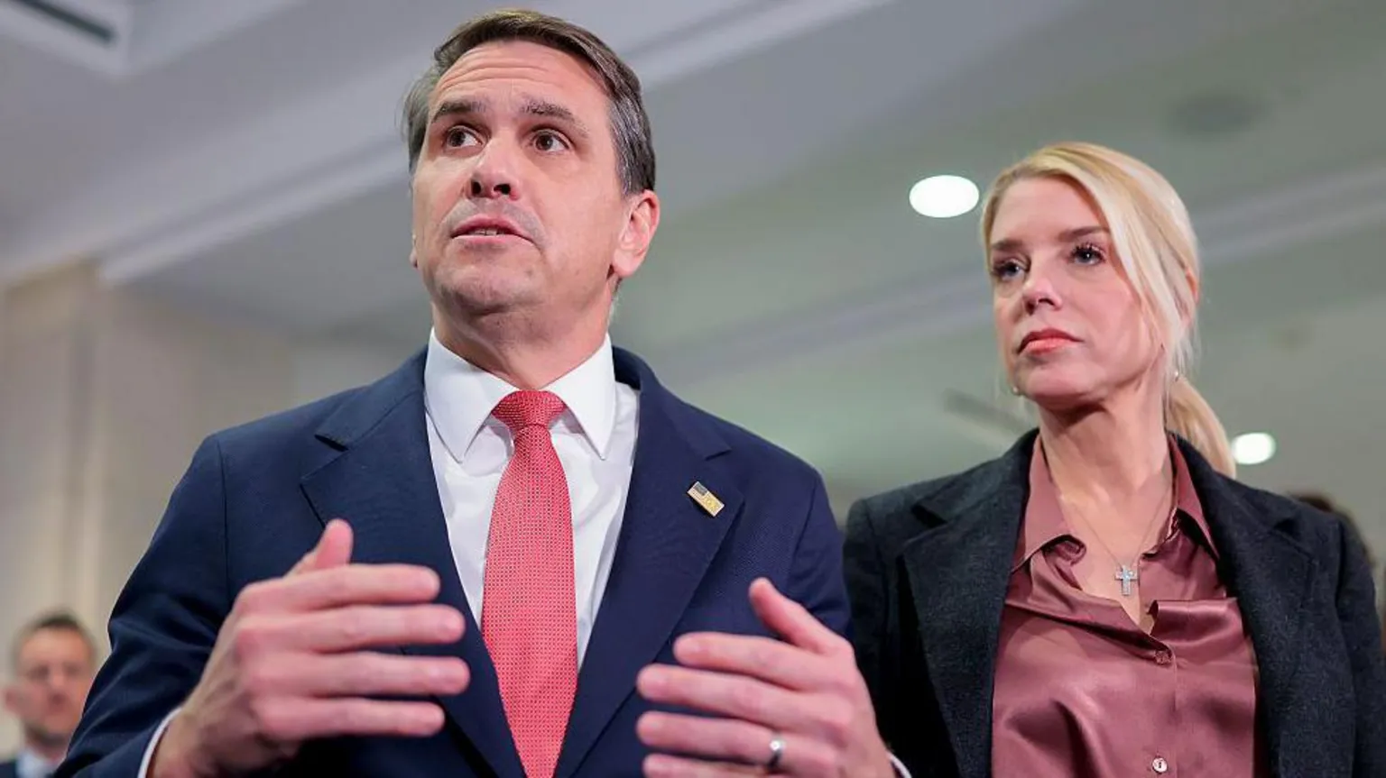  Todd Blanche and Pam Bondi speak with the media in the Capitol in March 2026. Blanche is wearing a blue suit and red tie, while Bondi is wearing a pink shirt and a black jacket 