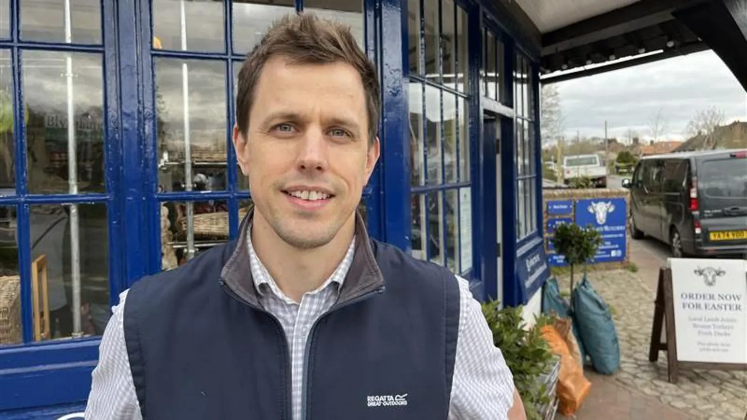 BBC/Phil Harrison A man with brown hair, blue gilet and check shirt stands in front of a shop with blue window frames. 