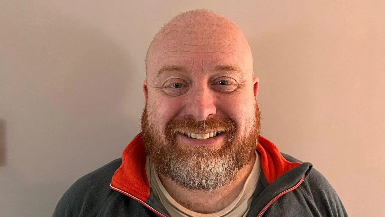 Jamie Lewis Jamie Lewis smiles at the camera showing his teeth. He is bald and has a ginger beard and blue eyes. He wears a grey sweatshirt and stands in front of a wall. 